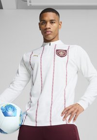 Baltas ilgas sportinis džemperis su raudonomis vertikaliomis juostelėmis, aukštu apykakle ir Menčesterio miesto logotipu. Modelis laiko mėlynai baltą futbolo kamuolį.