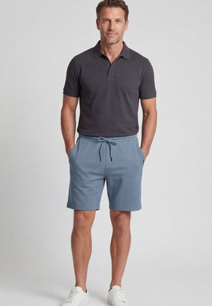 Mann steht mit den Händen in den Taschen blauer Shorts mit Kordelzug, trägt ein dunkles Poloshirt und weiße Sneakers vor einem einfarbigen Hintergrund.
