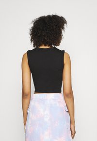 Žena s kudrnatými vlasy, oblečená v černém bezrukávovém crop topu a pastelové tie-dye sukni, stojící zády k prázdnému pozadí.