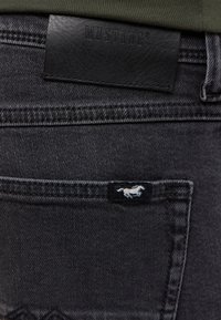 Schwarze Denim-Jeans mit einem geprägten Lederlabel mit der Aufschrift "MUSTANG" und einem kleinen schwarzen Anhänger mit einem weißen Pferdelogo auf der Gesäßtasche.