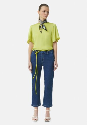 Bright lime green blouse with short sleeves, paired with dark blue denim cropped jeans featuring a knotted neon yellow belt. White strappy heels.