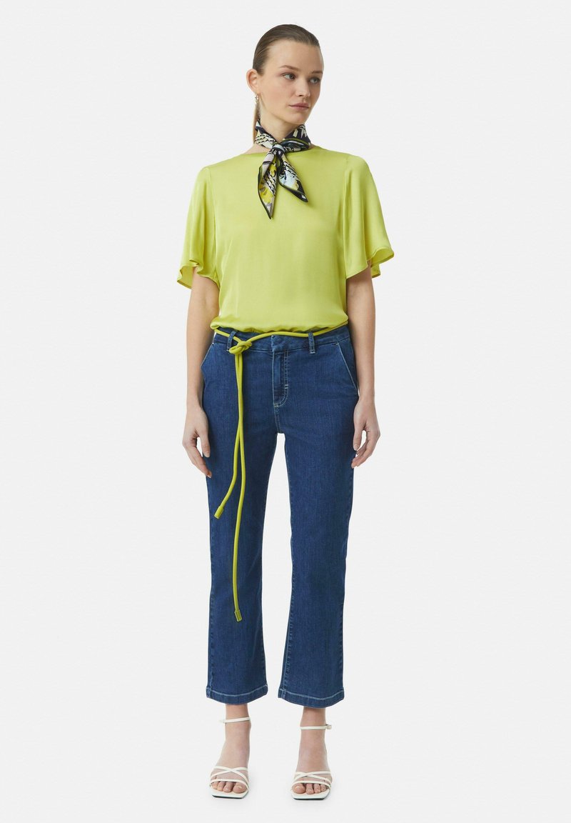 Bright lime green blouse with short sleeves, paired with dark blue denim cropped jeans featuring a knotted neon yellow belt. White strappy heels.