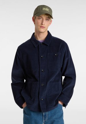 Marineblauwe corduroy jas met een puntige kraag, knoopsluiting, twee voorkepers en een gestructureerde stof; gecombineerd met een groene pet.