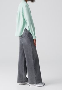 Camicia verde menta a maniche lunghe abbinata a jeans grigi a gamba larga. La camicia presenta una vestibilità comoda e polsini arricciati, mentre i jeans hanno una texture scolorita.