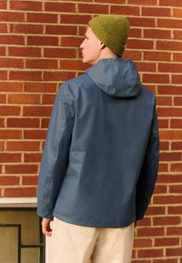 Veste imperméable bleue avec capuche, dotée d'une texture lisse et d'un design minimaliste. Associée à un bonnet en olive à côtes.