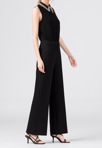 Svart ärmlös jumpsuit med pärlkrage och vid ben-design, med en figursydd midja och slät textur. Svarta rem-sandaler.