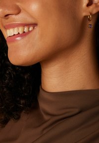 Goldene Creole mit einem kleinen lila Edelstein-Anhänger. Glatte Textur, kreisförmiges Design. Sanftes Licht reflektiert auf der goldenen Oberfläche.