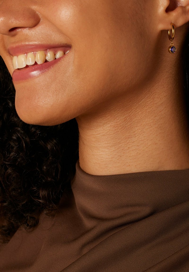 Goldene Creole mit einem kleinen lila Edelstein-Anhänger. Glatte Textur, kreisförmiges Design. Sanftes Licht reflektiert auf der goldenen Oberfläche.