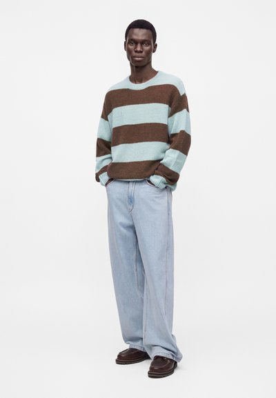 Homme portant un pull rayé bleu et marron, un jean large bleu clair et des chaussures marron foncé, debout les mains dans les poches.