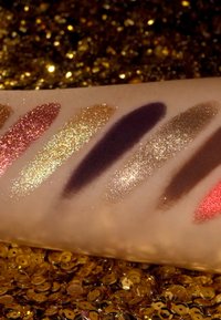 Campioni di otto tonalità cosmetiche su una superficie chiara, con colori come rosa, oro, viola scuro e rosso, con finiture scintillanti.