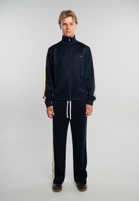 Hombre joven de pie mirando hacia adelante, vistiendo un chándal azul marino con franjas laterales crema y zapatos marrón oscuro, sobre un fondo claro y liso.