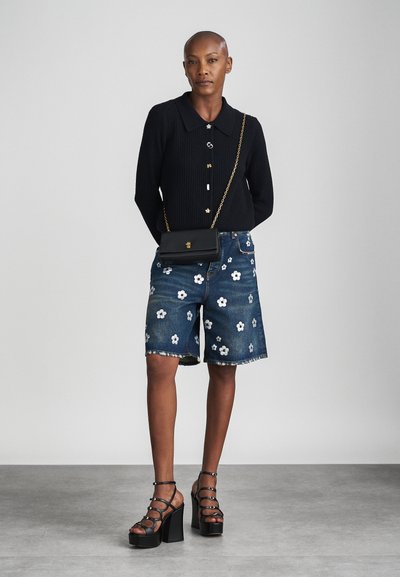 Persoon die een zwarte cardigan met knopen draagt, spijkershorts met witte bloemenprints, zwarte sandalen met plateauzolen en bandjes, en een kleine zwarte tas met gouden ketting.