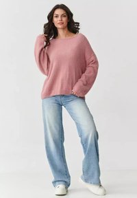 Jasnoróżowy oversize'owy sweter o fakturowym materiale, zestawiony z jasnobłękitnymi spodniami o szerokich nogawkach i białymi trampkami, na jednolitym tle.