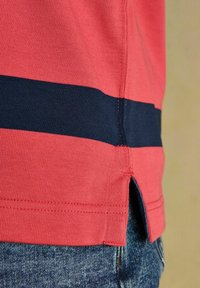 Punainen neulottu paita, jossa on vaakasuora tummansininen raita helmassa ja sivuhalkio, käytetty sinisten farkkujen päällä.