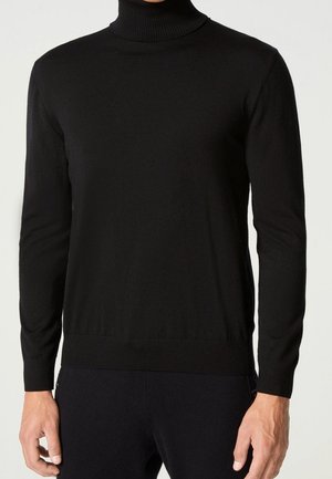 Man wearing a fitted black turtleneck sweater with ribbed cuffs and hem, standing against a plain light background.