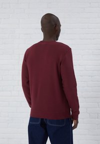 Felpa bordeaux con maniche lunghe, scollatura a girocollo e polsini a coste. Accoppiata con jeans in denim blu scuro su uno sfondo di mattoni bianchi.