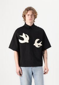 Camisa preta de manga curta feita de algodão, com dois silhuetas de pássaros em creme bordadas no peito e um fecho com botões à frente.