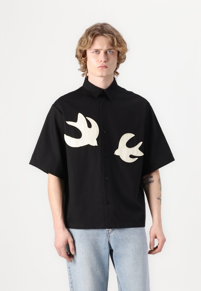 Schwarzes Kurzarmhemd aus Baumwolle, mit zwei cremefarbenen Vogelsilhouetten auf der Brust gestickt und einer Knopfleiste vorne.