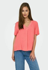 Koraalroze blouse met V-hals, korte mouwen, textuurpuntjes over de hele blouse en kantdetails bij de halslijn. Draag het met een blauwe jeans.