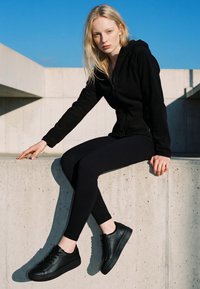 Mlada žena u crnoj trenirci s kapuljačom, tajicama i tenisicama sjedi na betonskoj ogradi nasuprot vedrom plavom nebu.