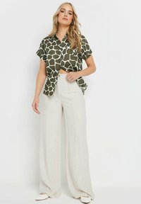 Camicia verde a maniche corte con un motivo geometrico bianco, abbinata a pantaloni beige a gamba larga e scarpe bianche slip-on.