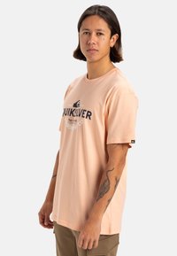 Camiseta de algodón de color melocotón, de manga corta, con un logotipo impreso en negro y en bold. El corte es holgado y cuenta con una etiqueta en la manga izquierda.