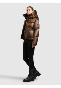 Brązowa kurtka puchowa z wysokim kołnierzem, błyszczącą fakturą, zamkiem z przodu i dwoma zamykanymi na zamek bocznymi kieszeniami. W połączeniu z czarnymi legginsami i botkami.