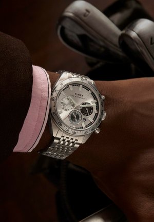 Silver stainless steel watch with chronograph features, white dial, black accents, and a woven band, worn on a wrist beside golf clubs.