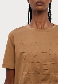 T-shirt in cotone marrone con un design in rilievo e testurizzato sulla parte anteriore. Collo a giro con maniche corte e finitura liscia.
