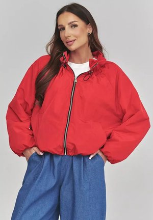 Kobieta ubrana w jasnoczerwoną kurtkę zapinaną na zamek z troczkiem przy kołnierzu i luźne niebieskie spodnie, pozująca z rękami w kieszeniach na jednolitym tle.