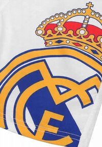 Biała tkanina z niebieskim i żółtym logo Realu Madryt, z czerwono-złotą koroną powyżej. Podkreśla wyraziste litery i gładką teksturę.