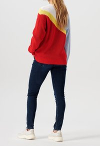 Nainen seisoo kädet taskuissa, päällään punainen, keltainen, valkoinen ja sininen villapaita, tummansiniset tiukat farkut ja valkoiset lenkkarit, selkä kameraan.