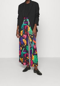 Personne portant un blazer noir, un haut noir et un pantalon large à motifs de fruits tropicaux colorés et de feuilles, associé à des mocassins noirs.