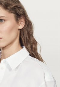 Camicia bianca con bottoni, collo strutturato e dettagli sulle spalle. Tessuto in cotone liscio, caratterizzata da un design minimalista e toni neutri.