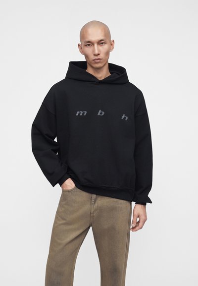 Homme portant un sweat à capuche noir, capuche baissée, avec les lettres grises m, b, h sur la poitrine, associé à un pantalon marron ample, debout avec une main dans la poche.