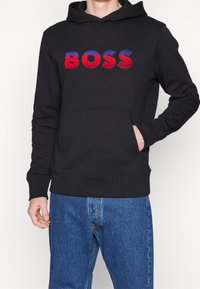 Felpa nera con cappuccio con logo "BOSS" rosso e blu, tasca frontale e polsini e orlo a costine, abbinata a jeans blu.