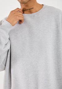 Felpa in cotone grigio chiaro, con una vestibilità ampia e colletto a coste. Presenta una texture leggera. Sotto il colletto, un tatuaggio recita "credi in te stesso."
