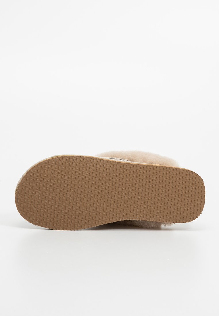 Beiger Hausschuh mit einem weichen, flauschigen Obermaterial und einer strukturierten braunen Gummisohle, die ein flaches Design und eine abgerundete Spitze ohne Verzierungen aufweist.