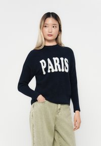 Námornícky sveter s bielym chlpatým nápisom "PARÍŽ". Má dlhé rukávy a okrúhly výstrih. Nosí sa s svetlozelenými nohavicami.