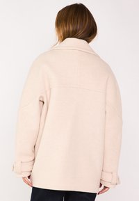 Cappotto oversize beige chiaro con colletto, spalle scese e polsini risvoltati. Tessuto morbido con una texture liscia e dettagli minimi.