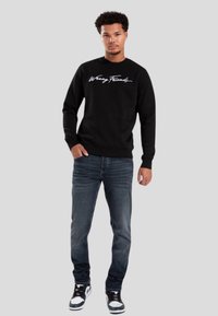 Sudadera negra con texto en cursiva blanca, combinada con jeans de mezclilla oscura y zapatillas en blanco y negro. Diseño sencillo, cuello redondo, mangas largas.