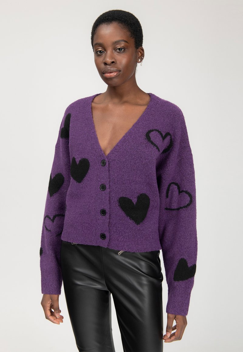 Una giovane donna indossa un cardigan viola con bottoni e motivi a cuore neri, insieme a pantaloni di pelle neri, mentre si trova di fronte a uno sfondo chiaro e semplice.