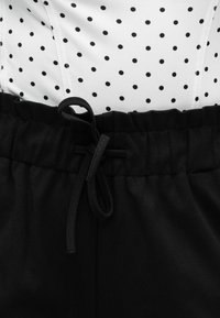 Pantalon à taille élastique noire avec un cordon de serrage noué, superposé sur un tissu blanc orné de pois noirs uniformément espacés.