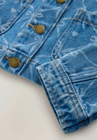 Close-up van mouw en knopen van een blauwe denimjack, met witte bloemmotieven en contrasterende oranje stiksel op de stof.