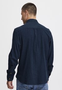 Temno modra dolgosleeve srajca iz teksturirane tkanine, ki ima klasičen ovratek, gumbnice na manšetah in podrobnost z naborkom na hrbtni strani.
