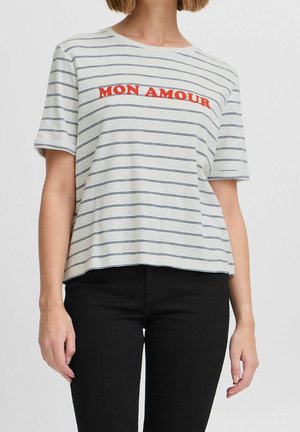 Vrouw draagt een wit T-shirt met korte mouwen met dunne zwarte horizontale strepen en rode tekst "MON AMOUR" over de borst, gecombineerd met een zwarte broek.