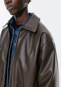 Hnedá umelá kožená bunda so štruktúrovaným golierom, s textúrovaným povrchom a zipsom, preložená cez modrú riflovú košeľu.