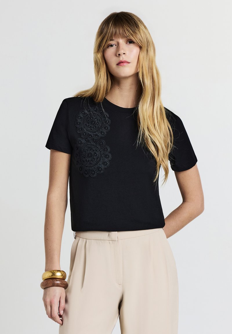 Donna con lunghi capelli biondi che indossa una t-shirt nera con dettagli in pizzo sulle spalle, pantaloni beige a vita alta e bracciali spessi in oro e legno.