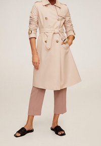 Žena v béžovém dvouřadém trenčkotě s páskem, s fialovými zkrácenými kalhotami a černými klouzavými sandály, stojící s jednou rukou v kapse.