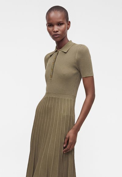 Femme portant une robe en maille côtelée vert olive avec des manches courtes, un col et des boutons sur le devant, debout devant un fond blanc uni.